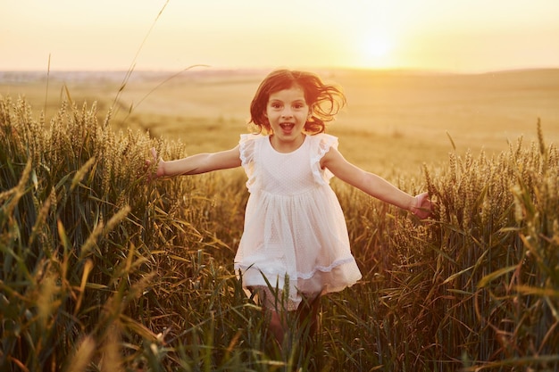 Bambina allegra in abito bianco che corre nel campo agricolo durante il giorno d'estate