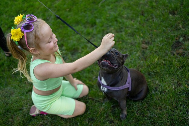 Bambina allegra in un vestito verde accanto a un cane di razza bulldog francese seduto sul prato nel parco