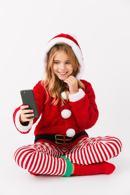 Bambina allegra sveglia che indossa il costume di Natale isolato, tenendo il telefono cellulare, prendendo un selfie