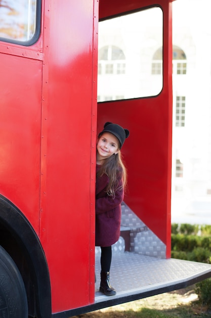 Bambina allegra vicino al bus inglese rosso in un bellissimo cappotto e un cappello. Viaggio del bambino. Scuolabus. Autobus rosso di Londra. Primavera. Con la giornata internazionale della donna. Dall'8 marzo!