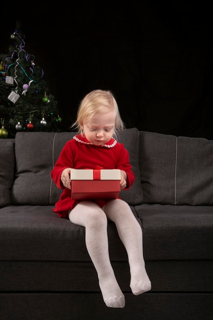 Bambina apre la confezione regalo seduto sul divano sullo sfondo di un albero di Natale Vigilia di Natale Regali di Capodanno Cornice verticale