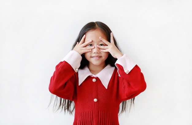 Bambina asiatica allegra che guarda tramite le sue mani su fondo bianco