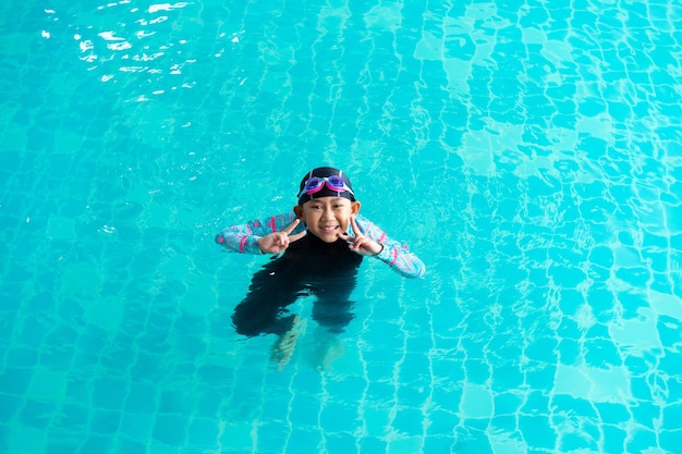 Bambina asiatica che indossa costume da bagno e berretto con occhiali sul bordo della piscina Impara a nuotare concetto di idea