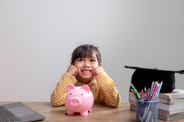 Bambina asiatica che indossa un cappello di graduazione con un salvadanaio rosa Risparmiare denaro investimento il futuro