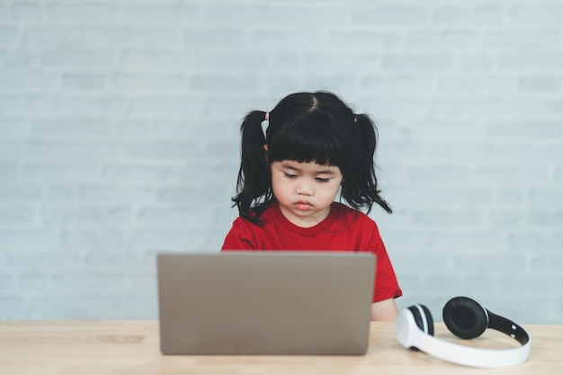 Bambina asiatica che indossa una maglietta rossa e una cuffia bianca usa il laptop e studia online sulla scrivania del tavolo in legno nel soggiorno di casa Istruzione che impara online dal concetto di casa