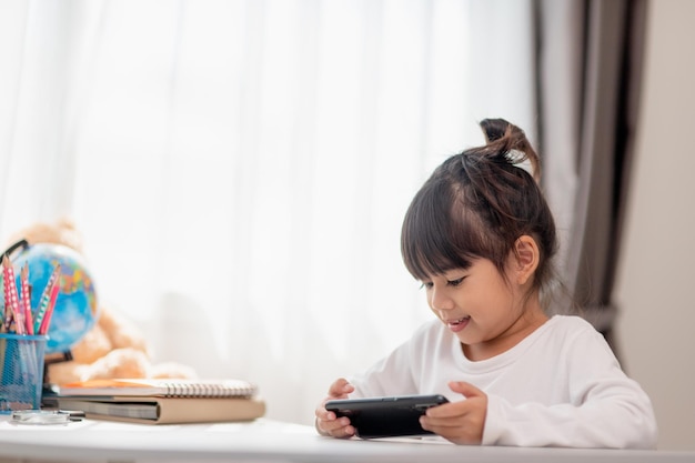 Bambina asiatica che utilizza un telefono da vicino distratto dallo studio seduto a un tavolo con i quaderni un bel bambino che si diverte con uno smartphone guardando il webinar homeschooling