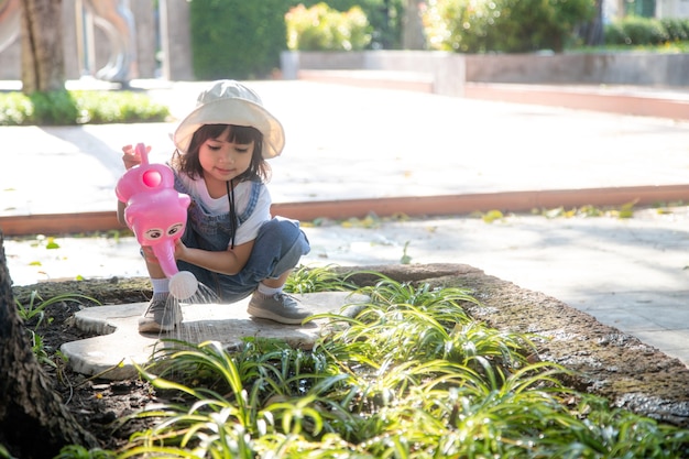 Bambina asiatica che versa acqua sugli alberi il bambino aiuta a prendersi cura delle piante con un annaffiatoio in giardino.