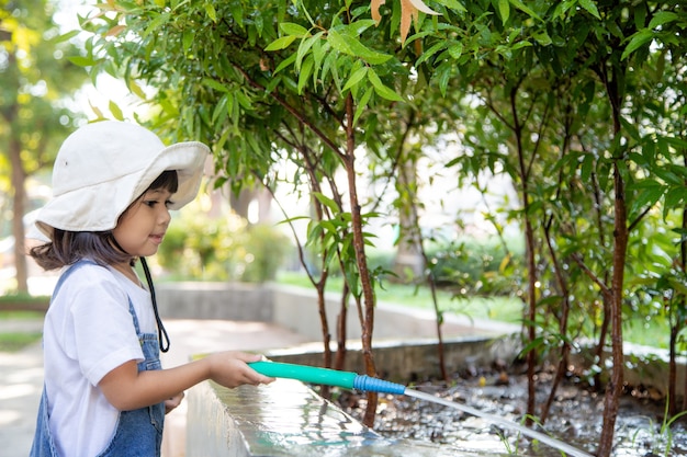 Bambina asiatica che versa acqua sugli alberi il bambino aiuta a prendersi cura delle piante con un annaffiatoio in giardino.