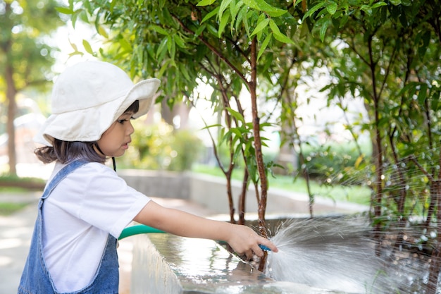 Bambina asiatica che versa acqua sugli alberi il bambino aiuta a prendersi cura delle piante con un annaffiatoio in giardino.