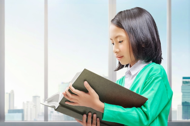 Bambina asiatica in piedi e leggendo il libro in casa. Torna al concetto di scuola.