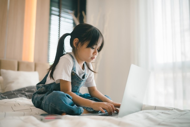 Bambina asiatica seduta a letto e che impara online sul laptop Internet Lezione di classe virtuale su video durante l'autoisolamento a casa Educazione video remota a distanza Studio scolastico moderno per bambini