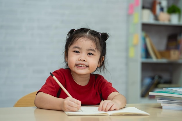 Bambina asiatica sorridente che indossa una camicia rossa scrive appunti in quaderno e libro di lettura per studiare online su tavolo di legno in salotto a casa Educazione apprendimento online da casa concetto