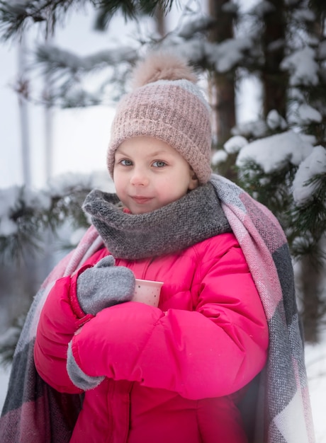 Bambina avvolta in una coperta che beve il tè nella foresta invernale