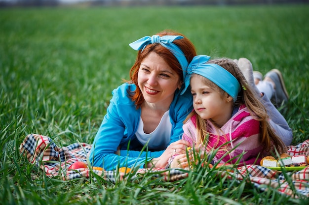 Bambina bambino e madre donna giacciono sul copriletto, erba verde nel campo, tempo primaverile soleggiato, sorriso e gioia del bambino, cielo blu con nuvole