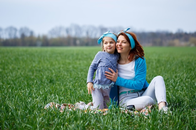 Bambina bambino e madre donna si siedono sul copriletto, erba verde nel campo, tempo soleggiato primaverile, sorriso e gioia del bambino, cielo blu con nuvole