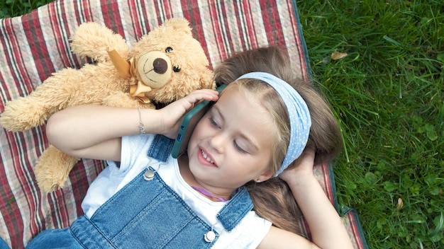 Bambina bambino felice posa sul prato verde con il suo orsacchiotto parlando al telefono cellulare.