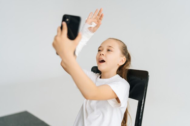 Bambina bionda carina con il braccio rotto avvolto in una benda di gesso bianco che parla di videochiamata su smartphone seduto in poltrona su sfondo bianco isolato