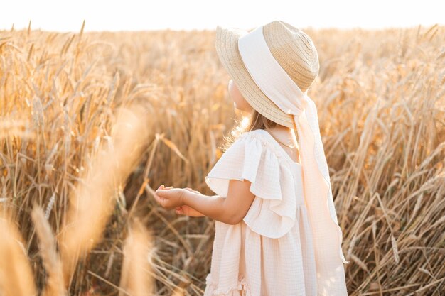 bambina bionda con cappello di paglia e abito di mussola beige nel campo di grano al tramonto