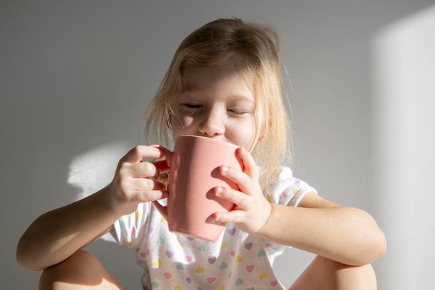 Bambina bionda con gli occhi chiusi tiene una tazza sullo sfondo di un muro bianco