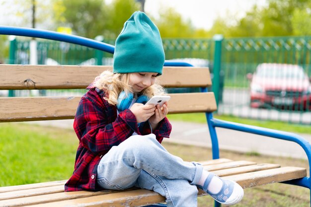 Bambina bionda con le mani e le cuffie dello smartphone, seduta su una panchina nel parco.