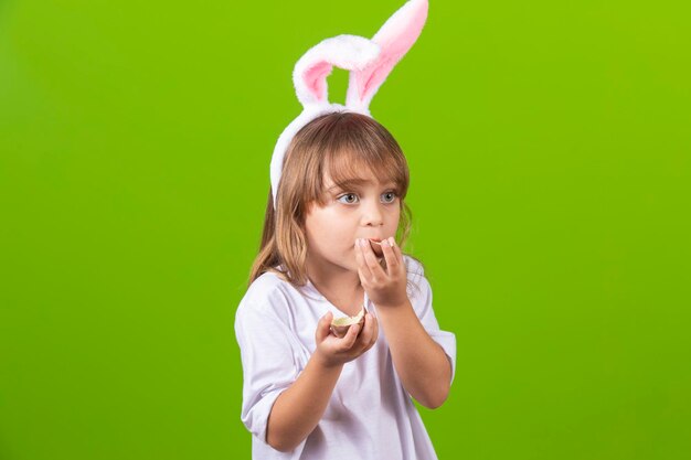 Bambina bionda con le orecchie di coniglietto di cioccolato sporche che mangiano l'uovo di Pasqua