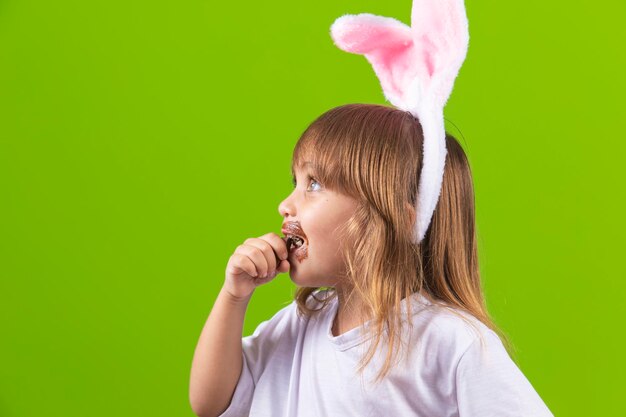 Bambina bionda con le orecchie di coniglietto di cioccolato sporche che mangiano l'uovo di Pasqua