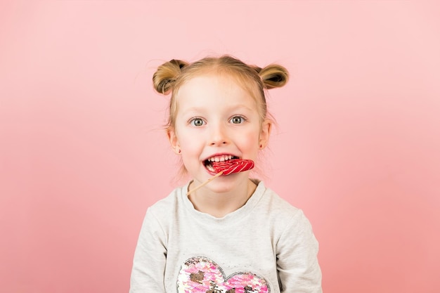 Bambina bionda divertente che sorride e gioca con il caramello lecca-lecca a forma di cuore rosso su sfondo rosa. Concetto di San Valentino o festa della mamma.