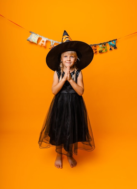 Bambina bionda in costume nero di halloween che ride e guarda la telecamera isolata su sfondo giallo