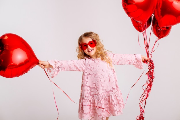 bambina bionda in un abito rosa è sorridente e in possesso di un sacco di palloncini a forma di cuore rosso concetto di San Valentino