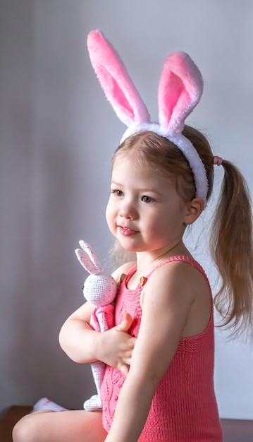 Bambina bionda in un pagliaccetto rosa con le orecchie del coniglietto di Pasqua sulla testa che abbraccia un coniglietto giocattolo