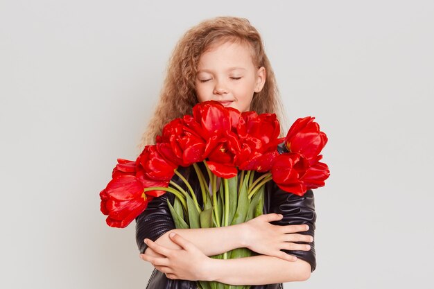 Bambina bionda sognante felice che abbraccia un sacco di tulipani rossi e tiene gli occhi chiusi, godendo del bellissimo presente, indossa una giacca nera