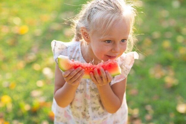 Bambina bionda sveglia che mangia anguria sull'erba nel parco