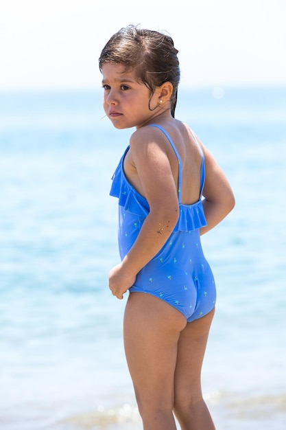 Bambina bruna che indossa un costume da bagno sulla spiaggia dalla parte posteriore di fronte all'oceano con il cielo sullo sfondo Concetto di vacanza al mare protezione solare mare stile di vita e relax