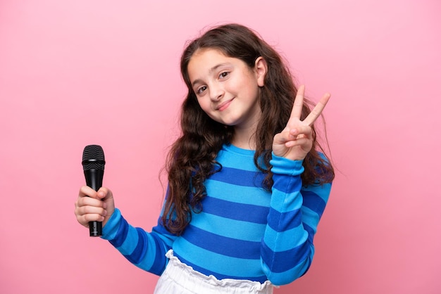 Bambina cantante che raccoglie un microfono isolato su sfondo rosa sorridente e mostra il segno di vittoria
