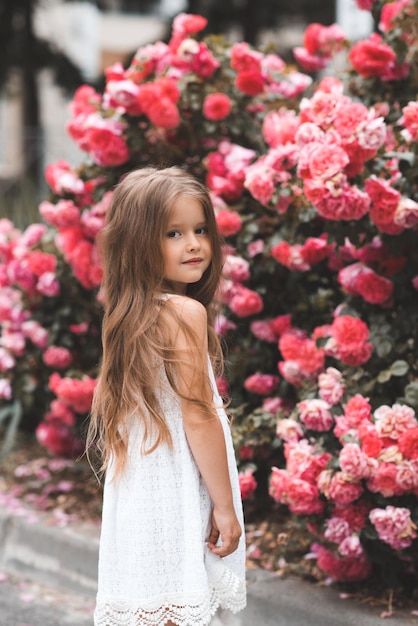 Bambina carina 45 anni con lunghi capelli biondi in posa su fiori rosa rosa sullo sfondo primo piano Primavera Bambino sorridente su cespugli fioriti in giardino Stagione estiva Felicità