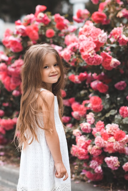 Bambina carina 45 anni con lunghi capelli biondi in posa su fiori rosa rosa sullo sfondo primo piano Primavera Bambino sorridente su cespugli fioriti in giardino Stagione estiva Felicità