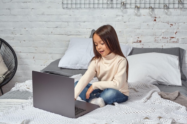 Bambina carina che si sente divertente mentre guarda i cartoni animati su un laptop seduta sul letto