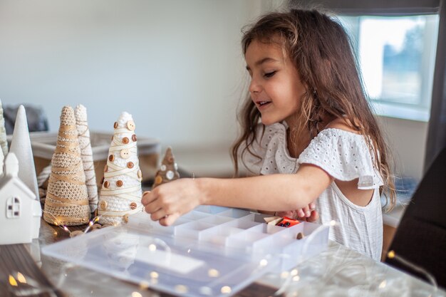 Bambina carina con i capelli ricci che fa mestieri e decora il cono dell'albero di Natale con i bottoni