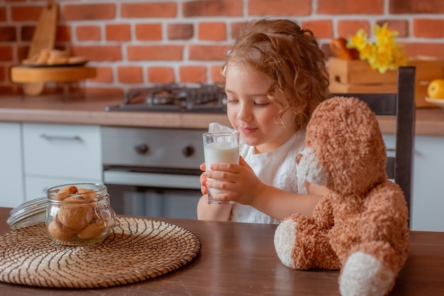 Bambina carina con orsacchiotto che beve latte per colazione in cucina a casa