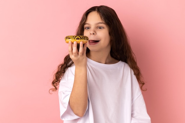 Bambina carina contentissima che lecca deliziosa ciambella guardando con desiderio di mangiare un dessert dolce