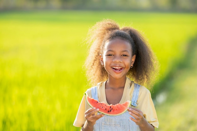 Bambina carina dalla pelle nera che mangia anguria all'aperto sullo sfondo del campo di riso verde Bambino africano che mangia anguria