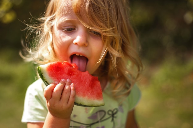 Bambina carina e divertente che mangia un pezzo di anguria in giardino in estate