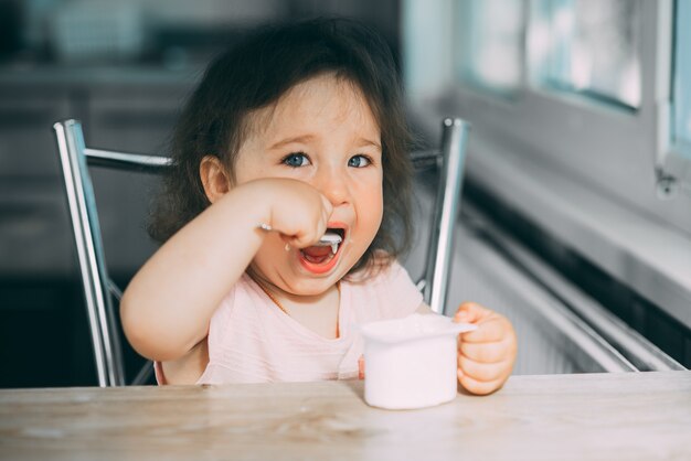 Bambina carina e divertente che mangia yogurt in cucina in un vestito rosa nel pomeriggio carino