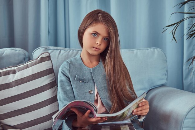 Bambina carina ed elegante con lunghi capelli castani, seduta su un divano, tiene diari di moda.