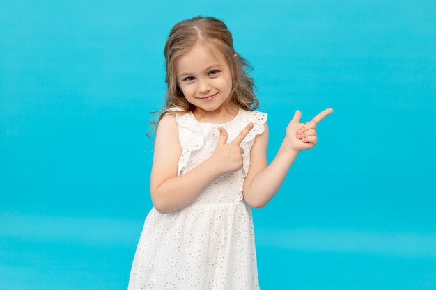 Bambina carina felice in un vestito bianco di cotone su sfondo blu in studio ridendo mostrando le dita nel ver sorridente e scherzando in un posto per il testo