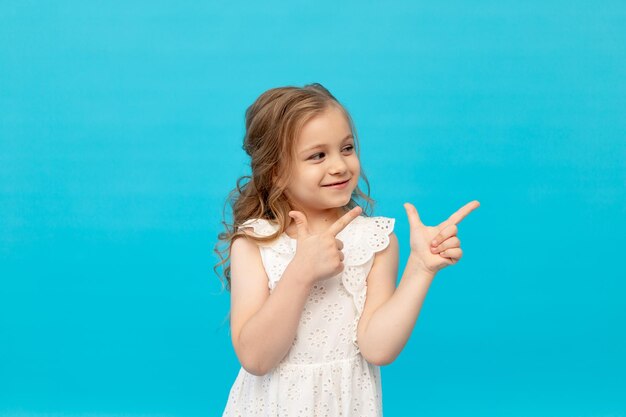 Bambina carina felice in un vestito bianco di cotone su sfondo blu in studio ridendo mostrando le dita nel ver sorridente e scherzando in un posto per il testo