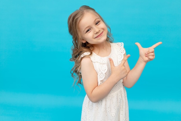 Bambina carina felice in un vestito bianco di cotone su sfondo blu in studio ridendo mostrando le dita nel ver sorridente e scherzando in un posto per il testo