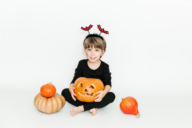 Bambina carina in abiti neri e cerchio di pipistrello. Tra zucche. Festa di Halloween. Sfondo bianco. Stile minimalista
