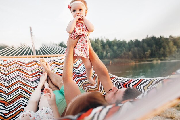 Bambina carina in abito elegante con i suoi genitori sdraiati su un'amaca vicino al lago e alla foresta durante un viaggio di vacanza in famiglia adorabile famiglia hipster con figlia