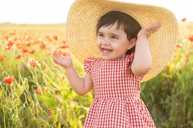 bambina carina in abito rosso e cappello di paglia sul campo di papaveri al tramonto estivo